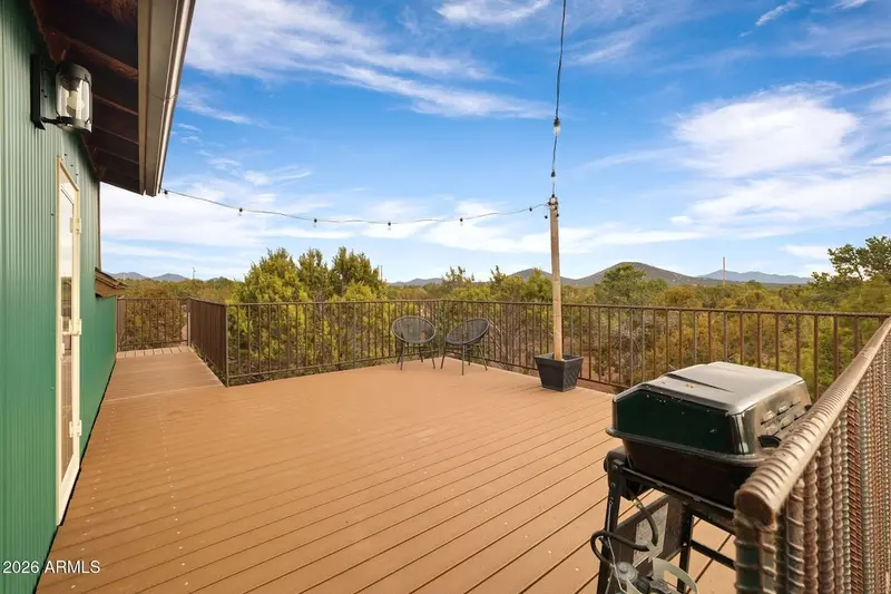 The Trex deck in daylight: green ribbed siding and a wall lantern at the left, string lights strung from a wooden post, black metal railing, two round wire chairs with a small table, and a covered gas grill at the right, with juniper forest and low mountains beyond.