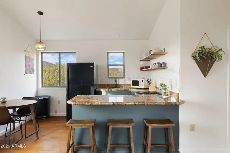 The kitchen seen across the dining area: a mottled granite peninsula with three wooden saddle stools, teal-blue cabinets, a black refrigerator, gas range and microwave, white tile backsplash, open wood shelves, a hanging plant, and a round table by a window looking into the trees.