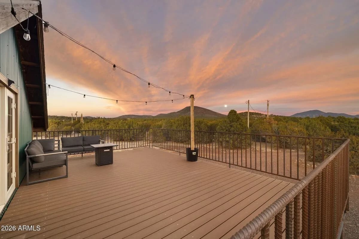 The wrap-around deck at sunset: grey outdoor sofas around a fire-pit table, string lights strung overhead, and the railing looking out over juniper forest to a dark hill under a pink and orange sky.