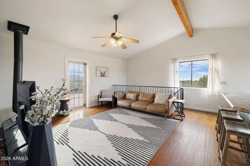 An open living area under a vaulted white ceiling with an exposed wood beam: a black free-standing stove and a vase of white blossom at the left, a tan leather sofa against a black metal stair rail, an armchair, a glazed door out to the deck, and a granite bar with stools at the right.