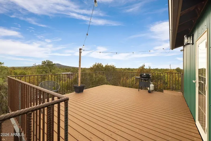 The deck looking along its length on a clear day: wide Trex boards, black railing, a wooden post carrying string lights, a potted plant, and a black gas grill with a propane tank standing against the green ribbed wall and its glazed door, juniper forest and a low hill beyond.