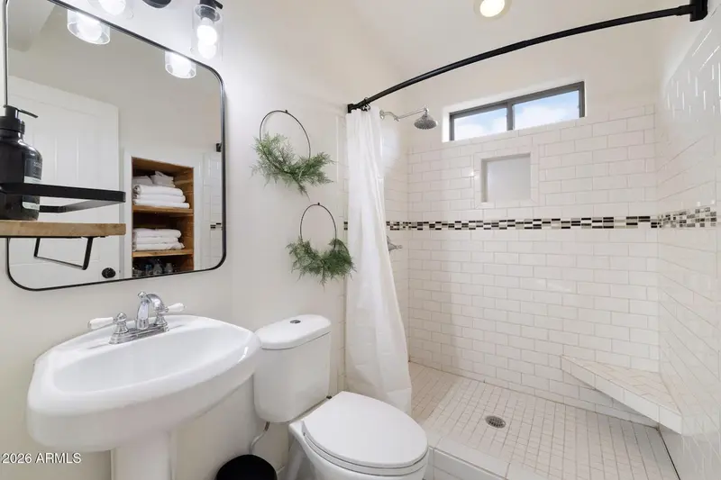 A bathroom with a pedestal basin, a black-framed mirror and vanity lights, a black wood shelf of rolled towels reflected in the mirror, two hanging greenery wreaths, and a walk-in shower tiled in white subway tile with a dark mosaic stripe under a small high window.