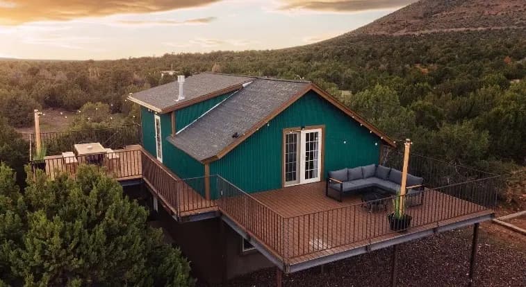 The cabin from the air at dusk: a green ribbed metal upper storey under a shingled gable roof, decking wrapping it on two levels with an outdoor sectional on one side and a table and chairs on the other, juniper forest all around and a long mesa behind, the horizon still lit.