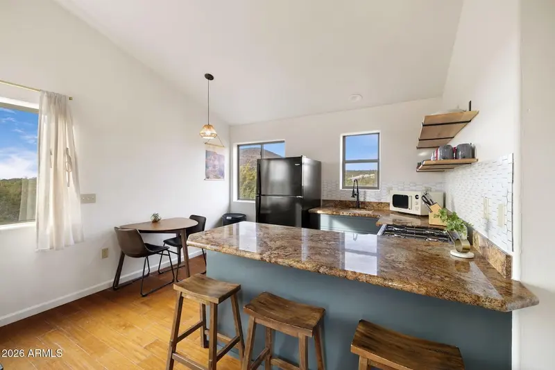 The kitchen and its eat-in corner: a granite peninsula with wooden saddle stools, teal cabinets, a black refrigerator, gas range and microwave, open shelves of mugs, and a pendant light over a small round table with two chairs beside a curtained window.