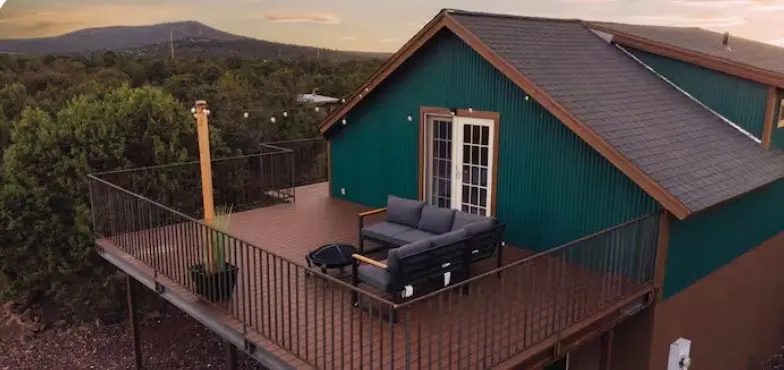The cabin from the air at dusk, closer in: the green ribbed wall and its glazed doors, a grey sectional and a fire-pit table on the decking, a wooden post carrying string lights, black railing, and juniper forest running out to a long mesa under a pale sunset.