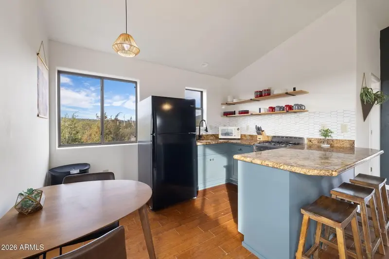 The kitchen and dining area from the doorway: teal-blue lower cabinets under mottled granite counters, a black refrigerator, gas range and microwave, open wood shelves holding red and grey mugs, a large window over the dining table, and saddle stools drawn up to the peninsula.