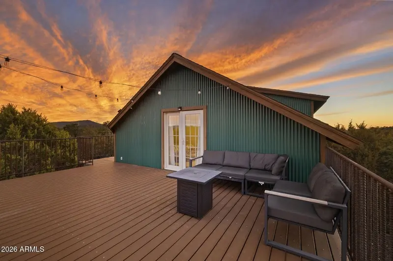 The deck at sunset facing the gable end: green ribbed siding with a glazed door, a grey sectional and a fire-pit table, a second grey sofa, string lights overhead, and a sky of orange and blue cloud over the junipers.