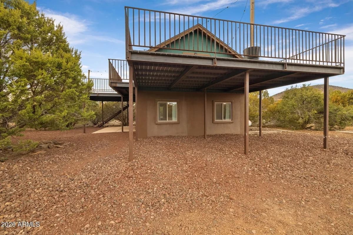 The cabin seen from underneath the deck: the stucco lower storey and its two windows, steel posts holding the deck overhead, wooden stairs at the side, and open gravel ground running out to the junipers.