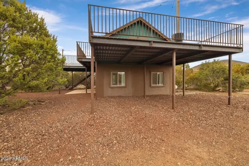 The cabin from underneath the deck: a stucco lower storey with two windows, steel posts carrying the deck overhead, stairs at the left, the green gable above, and gravel ground running out to junipers under a blue sky.