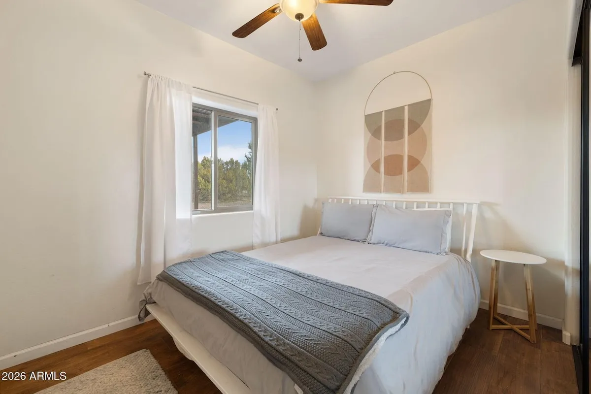 A main-level bedroom: a bed with white linen and a grey knit throw under a ceiling fan, a curtained window looking out to junipers, and a small round side table.