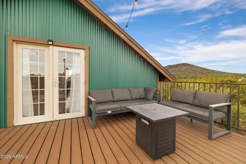 The deck against the green ribbed gable wall: a pair of glazed doors in a wood surround, a grey sectional and a second grey sofa around a fire-pit table, string lights overhead, and a rounded hill above juniper forest beyond the railing.