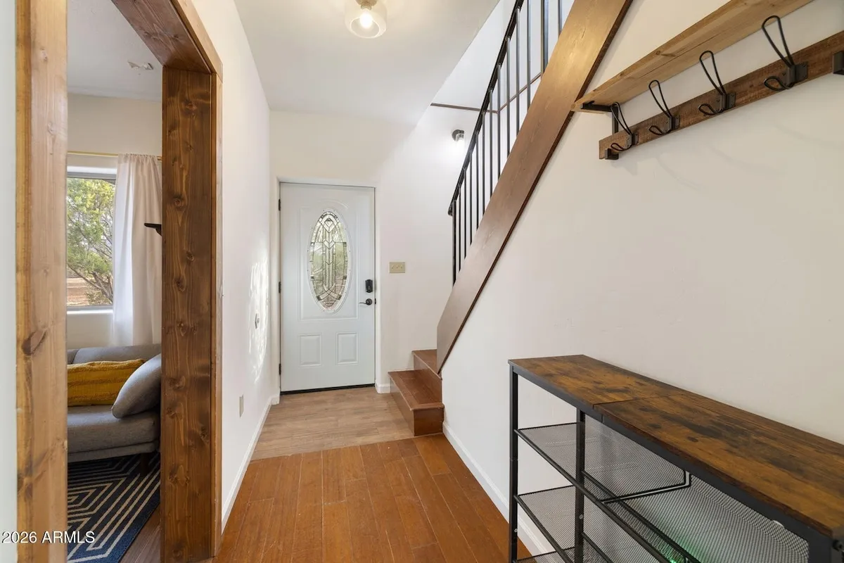 The entry hall: a white front door with an oval glass panel and a black keypad lock beside the handle, a wooden staircase rising to the right under a coat rail, and a doorway into the living area on the left.