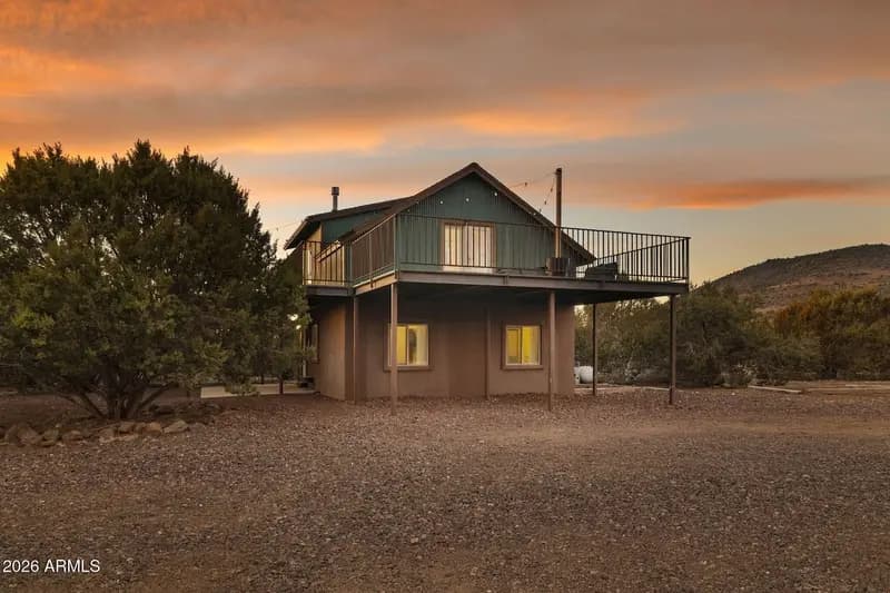 The cabin at dusk from across the gravel yard: the green upper storey and stucco base standing on steel posts, decking along the front, two windows lit yellow, a large juniper at the left and a hill at the right under an orange sky.
