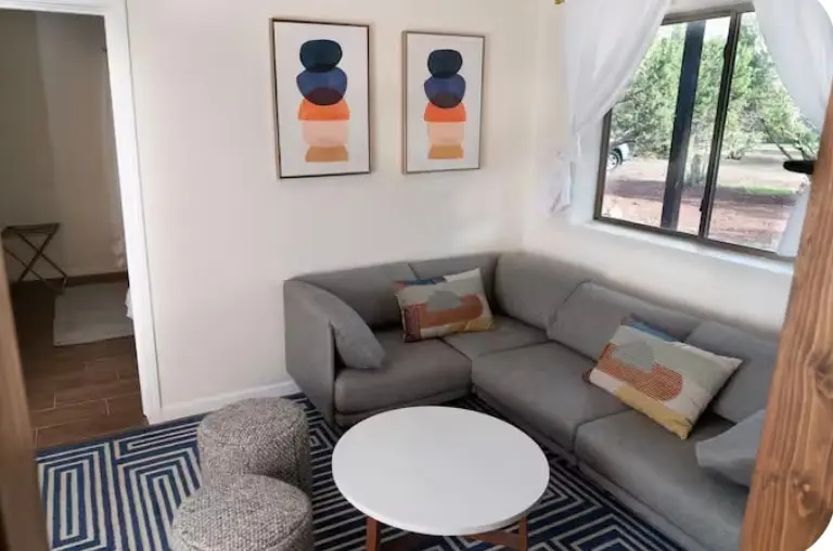 A small sitting room: a grey corner sofa with patterned cushions, two woven round poufs, a white oval coffee table on a blue and white striped rug, two abstract prints on the wall, and a window looking out to gravel and trees.