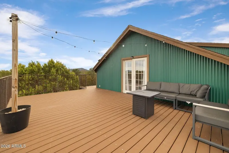 The deck in daylight looking toward the gable: wide Trex boards, a wooden post rising from a planter and carrying string lights, black railing, the green ribbed wall with its glazed doors, a grey sectional and a fire-pit table, junipers and a distant hill under blue sky.