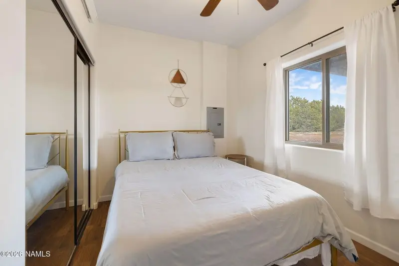 A bedroom with a brass-framed bed made up in white, mirrored closet doors reflecting it, a ceiling fan, a small hanging wall ornament, an electrical panel, and a curtained window looking out to trees and red earth.