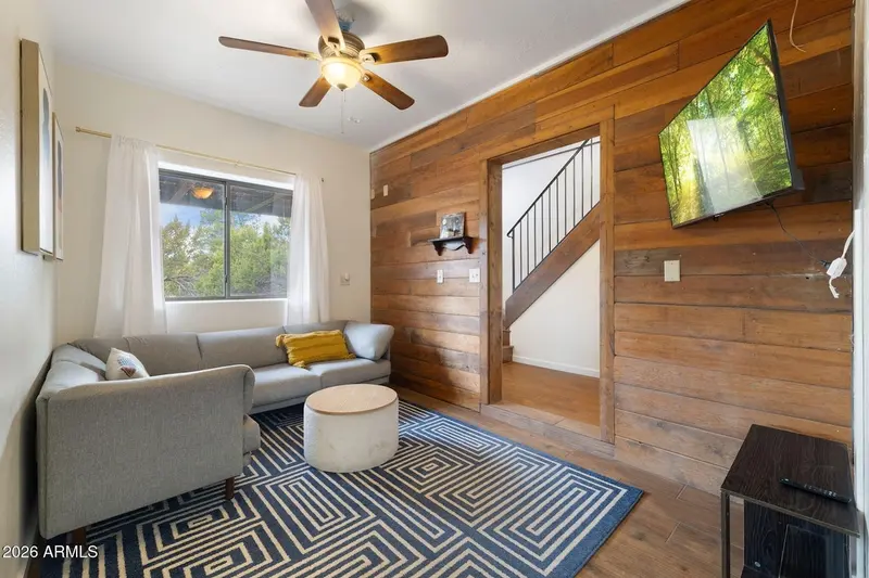 A living room with a reclaimed-wood plank wall carrying a wall-mounted television, an opening onto a white staircase, a grey sofa and armchair with a mustard cushion, a round upholstered ottoman, a blue geometric rug, and a window with junipers outside.