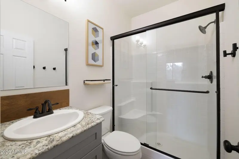 A bathroom with a granite-topped vanity over a grey cabinet, an oval basin and black tapware, a wood backsplash, a large frameless mirror, a framed geometric print, and a bath and shower behind dark-framed sliding glass doors.