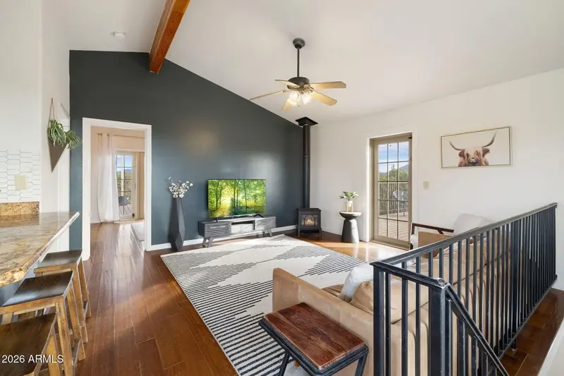 The living area seen over the stair rail: a vaulted ceiling with an exposed beam, a charcoal accent wall carrying a television and a black gas stove, a granite bar with stools at the left, a glazed door onto the deck, a longhorn print, and a leather sofa on a striped rug.
