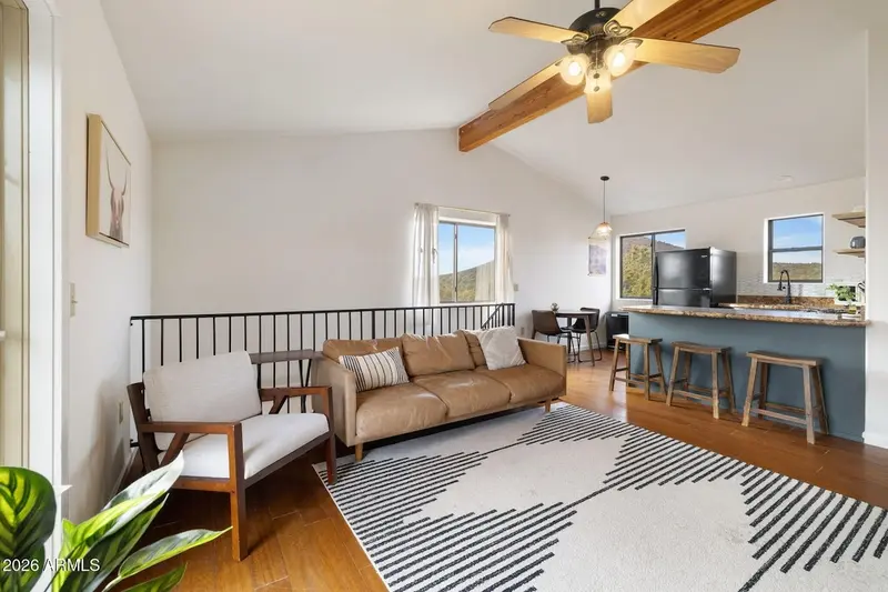 The living room looking toward the kitchen: a vaulted ceiling with an exposed beam and a brass fan, a tan leather sofa along the stair rail, a wood-framed armchair, a striped rug, windows onto the trees, and a teal-fronted bar with three wooden stools and a black refrigerator beyond.