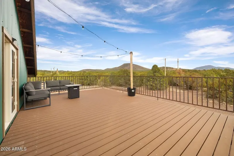 The open deck in daylight: a long run of Trex boards inside black railing, string lights on a wooden post, a grey sectional and fire-pit table at the far end against the green wall, and juniper forest with a distinct conical peak under a blue sky.
