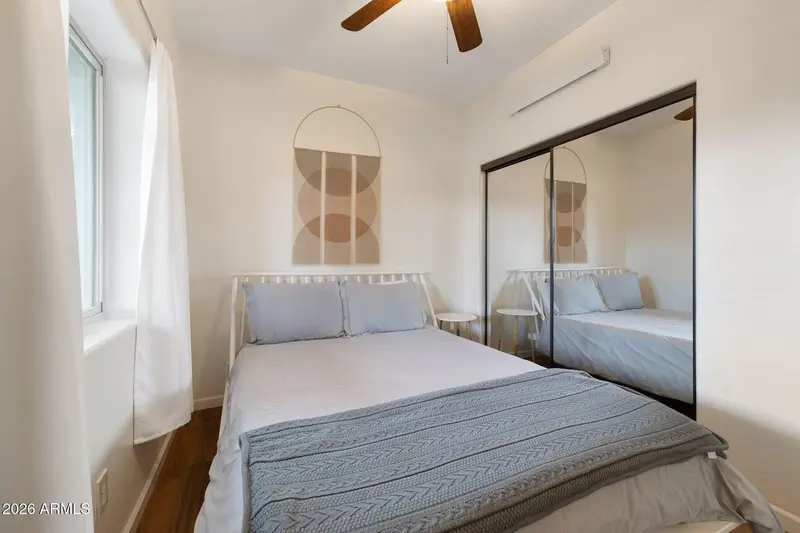 A bedroom from the foot of the bed: white bedding with a grey cable-knit throw, a slatted headboard, an arch-shaped print, mirrored closet doors filling the right-hand wall, a curtained window at the left, and a ceiling fan below a wall-mounted air handler.