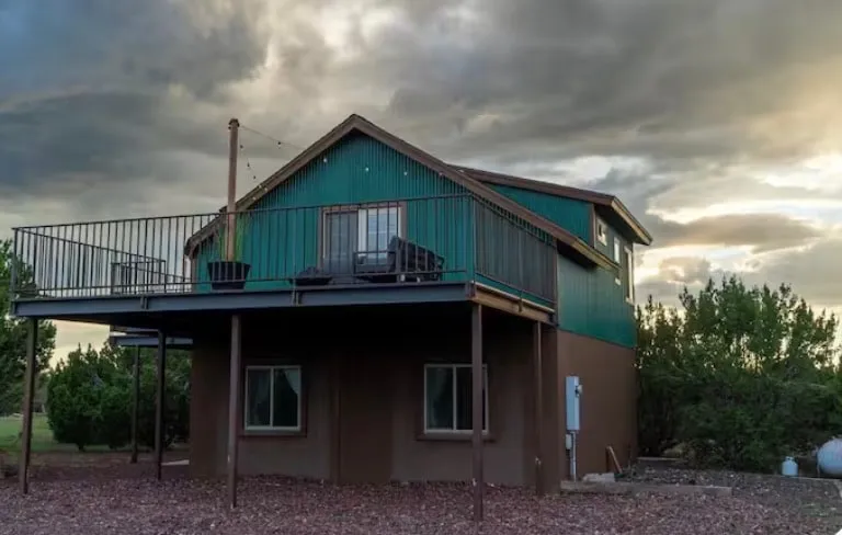 The cabin from the gravel yard: a green corrugated metal siding upper storey over a brown stucco base, the deck running along the upper floor on steel posts, junipers on both sides under a heavy gold-edged sky.