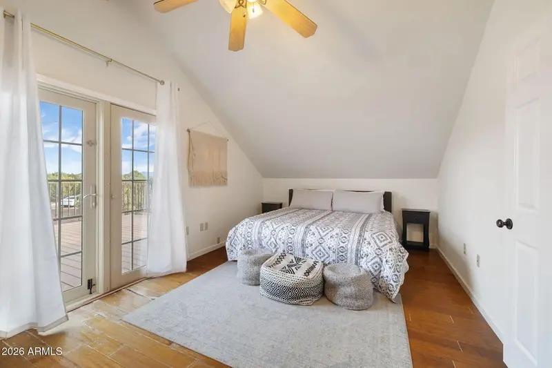 A bedroom under a sloping vaulted ceiling: a bed with a black and white patterned cover, three floor poufs at its foot, the centre one patterned and the two flanking it plain grey knit, curtained glazed doors opening onto the deck, a small stove-shaped heater, a woven wall hanging, and a grey rug on wood floor.
