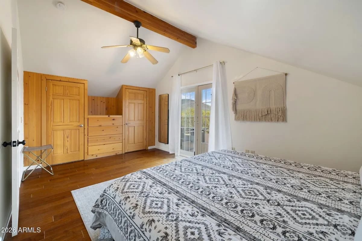 The upstairs primary bedroom: a large bed with a patterned cover under a vaulted ceiling and an exposed wood beam, pine wardrobes and a dresser, and French doors opening onto the deck.