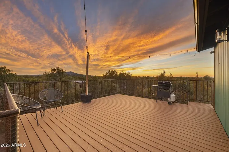 The deck at sunset: two round wire chairs at the left, a black gas grill with its propane tank at the right, black railing all around, a wooden post carrying string lights, and brilliant orange cloud spread over the junipers and a distant hill.