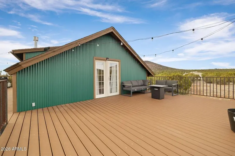 The deck in daylight from the far corner: the green ribbed gable wall with its glazed doors, a grey sectional and a fire-pit table set against it, string lights strung overhead, and a hill above juniper forest beyond the railing.