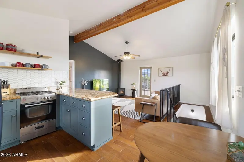 The kitchen looking through to the living area: a stainless gas range, teal cabinets under granite counters, white tile backsplash, open wood shelves of red and grey mugs, an exposed beam under the vault, the charcoal wall with its television and gas stove beyond, and a round dining table in the foreground.