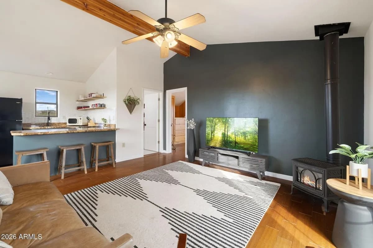 The open living space: a vaulted ceiling with an exposed beam, a granite breakfast bar with wooden stools and the kitchen behind it, a leather sofa on a striped rug, and a lit free-standing gas stove against a dark green wall.